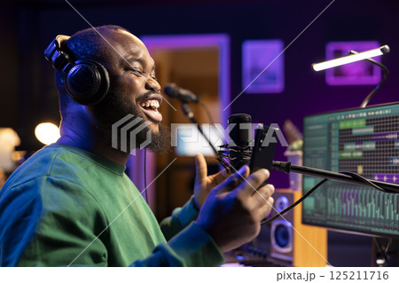 Songwriter producer recording a new track in home studio, singing on microphone to create an acoustic song. Artist singer performing soundtracks and reading lyrics on smartphone. 125211716