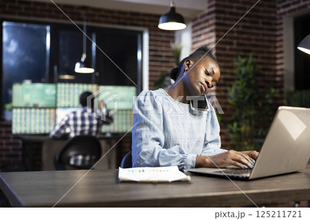 Black female employee manages multiple tasks at once, talking on her cellphone while typing financial data on laptop. African american woman using her personal computer and mobile device in office. 125211721
