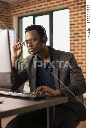 Pensive black man adjusting his glasses and working at desktop pc, focused on client information. Concentrated call center agent wearing headset, attending to customer support tasks on his computer. 125211735