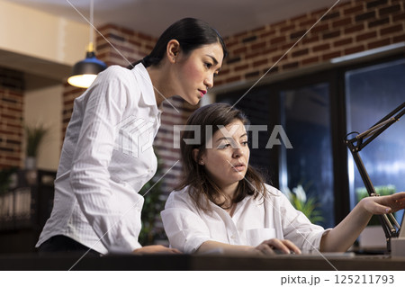 Dedicated female employees analyze research data on a laptop, having a business discussion. Asian manager stands and listens as her coworker explains the details, working together on a business task. 125211793