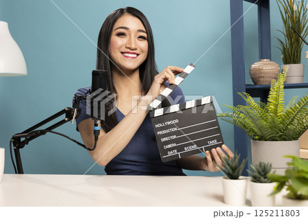 Female vlogger presents a clapperboard into the camera, sitting at a desk with a microphone. The scene highlights filmmaking equipment and atmosphere of video content creation for online reviews. Female vlogger presents a clapperboard into the camera, sitting at a desk with a microphone. The scene highlights filmmaking equipment and atmosphere of video content creation for online reviews. 125211803