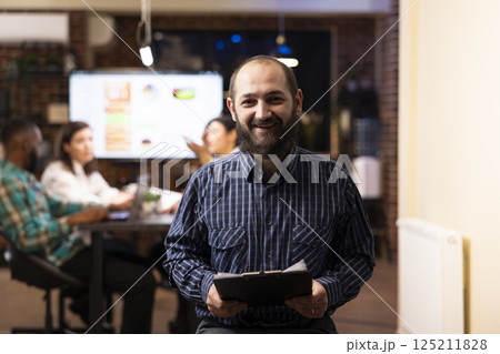 Caucasian male entrepreneur holds a clipboard in modern startup office. The night shift setting, brick wall and blurred team in background emphasize productivity and teamwork. 125211828