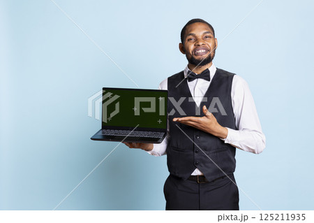 Front desk staff showing an isolated copy space on laptop in studio, wearing suit and tie greeting guests at resort. Professional bellhop working in hospitality industry and tourism. 125211935