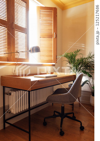 A close-up of a home workplace with a wooden table, a stylish gold lamp and a laptop. Bright sunlight from an open window with louvered shutters. A close-up of a home workplace with a wooden table, a stylish gold lamp and a laptop. Bright sunlight from an open window with louvered shutters. 125217686