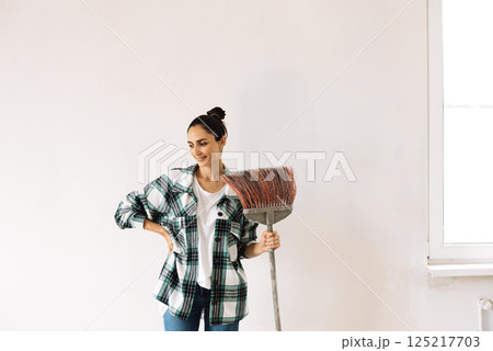 A young woman smiles and holds a sweeping brush against a white wall. A laughing girl in casual clothes is resting and holding a broom. A break between cleaning. 125217703