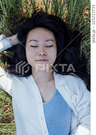 Asian woman in a white shirt and a gray top lies on the grass and laughs Asian woman in a white shirt and a gray top lies on the grass and laughs 125217733