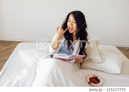 An attractive young Asian woman is lying in bed reading a book and eating berries. A charming female Korean is resting on the sofa in the bedroom. An attractive young Asian woman is lying in bed reading a book and eating berries. A charming female Korean is resting on the sofa in the bedroom. 125217735