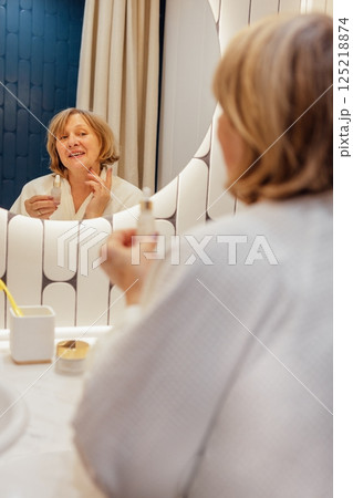 An elderly woman in a white terry dressing gown applies a serum to her face An elderly woman in a white terry dressing gown applies a serum to her face 125218874