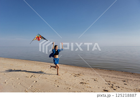A teenager in casual clothes flies a kite on the beach. The girl laughs and runs along the sandy beach. 125218887