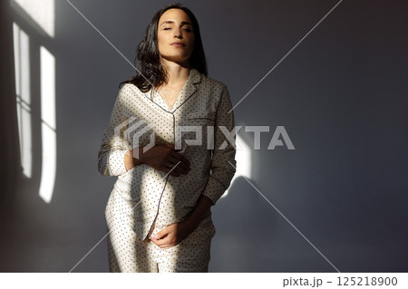 Pregnant woman stands in profile near window in dotted dress. Pregnant woman stands in profile near window in dotted dress. 125218900