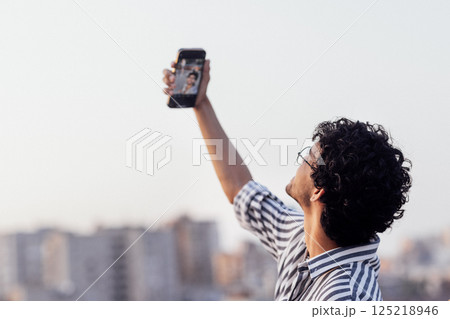 Young curly-haired guy in glasses and striped shirt answers video call on phone Young curly-haired guy in glasses and striped shirt answers video call on phone 125218946