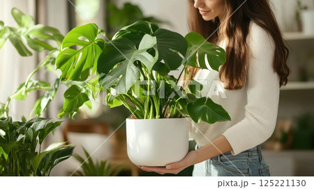 A woman in a cozy indoor setting carefully holds a lush potted monstera plant A woman in a cozy indoor setting carefully holds a lush potted monstera plant 125221130
