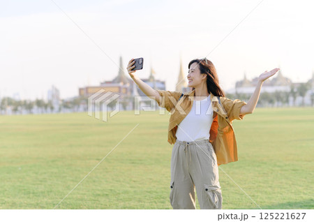 Traveler asian woman in her 30s making a livestream or selfie on smartphone while explores Wat Phra Kaew emerald Buddha. Share the wonders of Thai heritage through her journey. Traveler asian woman in her 30s making a livestream or selfie on smartphone while explores Wat Phra Kaew emerald Buddha. Share the wonders of Thai heritage through her journey. 125221627