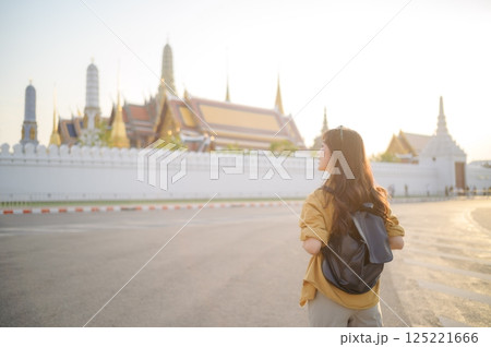 Traveler asian woman in her 30s, backpack slung over her shoulder, explores the intricate details of Wat Pra Kaew with childlike wonder. Sunlight dances on the golden rooftops. Traveler asian woman in her 30s, backpack slung over her shoulder, explores the intricate details of Wat Pra Kaew with childlike wonder. Sunlight dances on the golden rooftops. 125221666