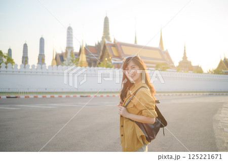 Traveler asian woman in her 30s, backpack slung over her shoulder, explores the intricate details of Wat Pra Kaew with childlike wonder. Sunlight dances on the golden rooftops. Traveler asian woman in her 30s, backpack slung over her shoulder, explores the intricate details of Wat Pra Kaew with childlike wonder. Sunlight dances on the golden rooftops. 125221671