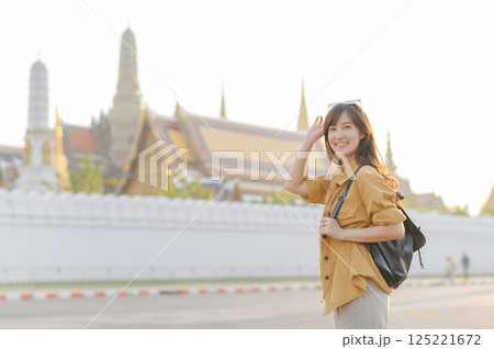 Traveler asian woman in her 30s, backpack slung over her shoulder, explores the intricate details of Wat Pra Kaew with childlike wonder. Sunlight dances on the golden rooftops. 125221672