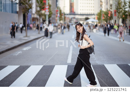 Urban joy unfolds as a young Asian woman navigates a busy crosswalk, seamlessly blending into the lively rhythm of local life. Urban joy unfolds as a young Asian woman navigates a busy crosswalk, seamlessly blending into the lively rhythm of local life. 125221741