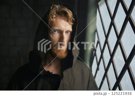 Portrait of pensive monk in medieval costume with hood looking through window of monastery 125221938