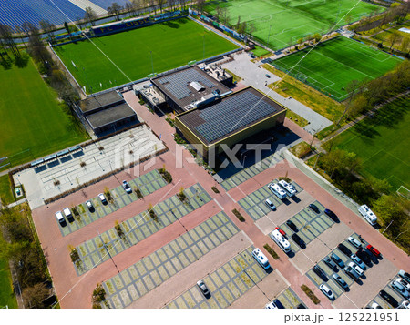 Aerial view of a sports complex with multiple football fields, a track and field stadium, tennis courts, and parking area, surrounded by lush forest and colorful tulip fields in the distance. Aerial view of a sports complex with multiple football fields, a track and field stadium, tennis courts, and parking area, surrounded by lush forest and colorful tulip fields in the distance. 125221951