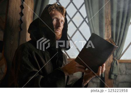 Old medieval monk reading sacred religious book praying with rosary beads in hand 125221958