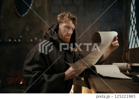 Medieval monk wearing robe with hood reading sacred manuscript on parchment Medieval monk wearing robe with hood reading sacred manuscript on parchment 125221965