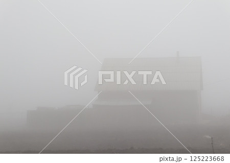 A captivating, mysterious foggy landscape featuring a rustic house that enhances the atmosphere A captivating, mysterious foggy landscape featuring a rustic house that enhances the atmosphere 125223668