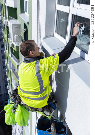 A Professional Window Cleaner in a Neon Yellow Shirt Safely Washing Windows on a High-Rise Building with a Suspended Harness and Equipment for Safety and Efficiency A Professional Window Cleaner in a Neon Yellow Shirt Safely Washing Windows on a High-Rise Building with a Suspended Harness and Equipment for Safety and Efficiency 125224891