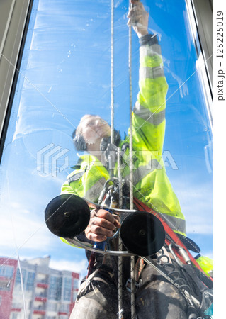 Professional Window Cleaner Man Cleans High-Rise Building Windows with Safety Harness and Equipment in Bright Blue Sky Environment Professional Window Cleaner Man Cleans High-Rise Building Windows with Safety Harness and Equipment in Bright Blue Sky Environment 125225019