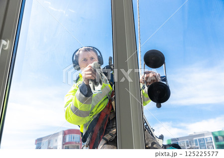 Professional Window Cleaner Performing Cleaning Task on High Rise Building with Reflections on Glass and Blue Sky Professional Window Cleaner Performing Cleaning Task on High Rise Building with Reflections on Glass and Blue Sky 125225577