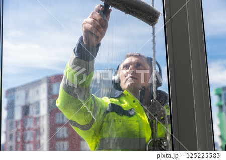 Professional Window Cleaner Performing His Task with Precision while Wearing Safety Gear on a Bright and Sunny Day, Enhancing the Appeal of High-Rise Buildings in the Background Professional Window Cleaner Performing His Task with Precision while Wearing Safety Gear on a Bright and Sunny Day, Enhancing the Appeal of High-Rise Buildings in the Background 125225583