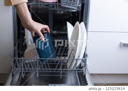 A Close-Up View of a Hand Reaching into a Dishwasher to Remove a Blue Textured Container Alongside Various Plates and Dishes Inside the Appliance 125225614