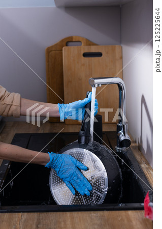 A Detailed View of a Person Cleaning a Pan Under Running Water in a Modern Kitchen Environment with Wooden Cutting Boards in the Background A Detailed View of a Person Cleaning a Pan Under Running Water in a Modern Kitchen Environment with Wooden Cutting Boards in the Background 125225644