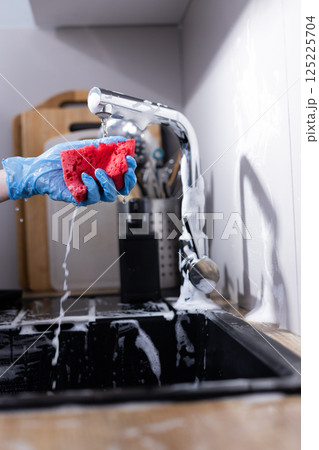 Bright Red Sponge Being Used to Clean a Sink in Modern Kitchen with Water and Soap Bubbles Flowing from the Faucet and Various Kitchen Utensils in the Background 125225704