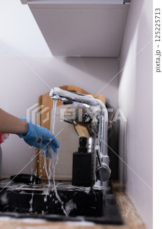 A close-up view of a hand using a faucet with soapy water, emphasizing cleanliness while preparing to do the dishes in a cozy kitchen setting with utensils in the background. 125225713