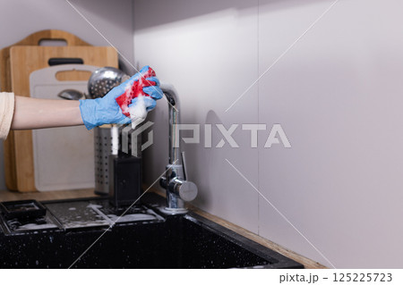 A Gloved Hand Holds a Colorful Sponge with Foamy Soap While Cleaning Dishes in a Modern Kitchen Setting Surrounded by Household Cleaning Supplies and a Neat Sink Area 125225723