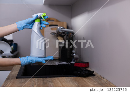 Hands in Blue Gloves Holding a Cleaning Spray Bottle in a Modern Kitchen Setting Focused on Hygiene and Cleanliness with Kitchen Utensils Visible in the Background. Hands in Blue Gloves Holding a Cleaning Spray Bottle in a Modern Kitchen Setting Focused on Hygiene and Cleanliness with Kitchen Utensils Visible in the Background. 125225736
