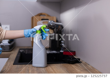 Hand in Blue Gloves Holding a Spray Bottle for Cleaning on a Wooden Countertop in a Modern Kitchen with a Sink and Kitchen Utensils in the Background Hand in Blue Gloves Holding a Spray Bottle for Cleaning on a Wooden Countertop in a Modern Kitchen with a Sink and Kitchen Utensils in the Background 125225737
