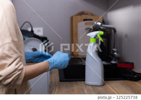 Essential Kitchen Cleaning Supplies: A Person in Blue Gloves Prepares to Use a Spray Bottle for Effective Disinfection on a Wooden Countertop 125225738