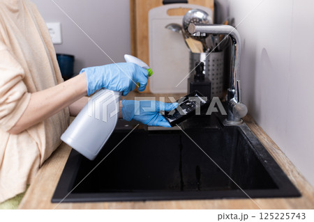 Person in Blue Gloves Cleaning a Black Sink with Spray Bottle and Dish Holder in a Modern Kitchen Setting, Emphasizing Household Cleaning Routine and Maintaining Hygiene Person in Blue Gloves Cleaning a Black Sink with Spray Bottle and Dish Holder in a Modern Kitchen Setting, Emphasizing Household Cleaning Routine and Maintaining Hygiene 125225743