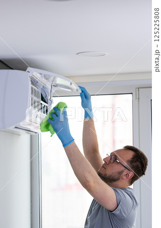 A Professional Wearing Blue Gloves Cleans the Indoor Unit of an Air Conditioner to Ensure Optimal Performance and Hygiene Inside a Modern Apartment in Bright Natural Lighting 125225808