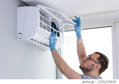 Professional Service: An Expert Technically Skilled Individual Reaches Up to Perform Maintenance on a Wall-Mounted Air Conditioning Unit for Optimal Performance and Longevity 125225816