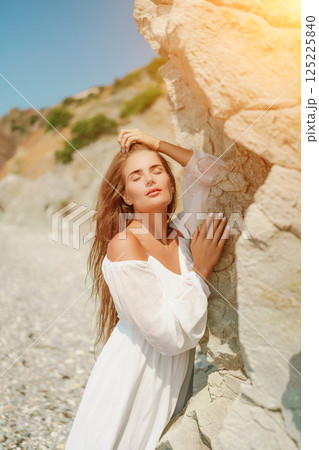 A woman in a white dress is standing on a rocky beach. Concept of serenity and tranquility, as the woman is enjoying the natural beauty of the beach. 125225840