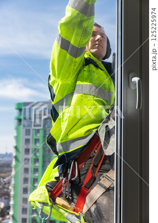 A Dedicated Window Cleaner in a Yellow Safety Suit Working on a High-Rise Building While Harnessed for Safety and Using Professional Equipment to Ensure a Flawless Shine A Dedicated Window Cleaner in a Yellow Safety Suit Working on a High-Rise Building While Harnessed for Safety and Using Professional Equipment to Ensure a Flawless Shine 125225974