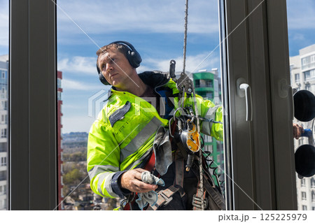 Skilled Window Cleaner Working on High-Rise Building, Captured in Bright Daylight, Wearing Safety Gear and Headphones for Enhanced Focus and Protection Skilled Window Cleaner Working on High-Rise Building, Captured in Bright Daylight, Wearing Safety Gear and Headphones for Enhanced Focus and Protection 125225979