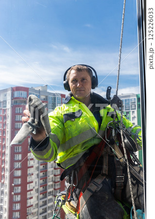 Professional Window Cleaner at Work: A Skilled Worker in High-Rise Building Wearing Safety Gear and Headphones While Cleaning Windows in a Bright Blue Sky Professional Window Cleaner at Work: A Skilled Worker in High-Rise Building Wearing Safety Gear and Headphones While Cleaning Windows in a Bright Blue Sky 125226033