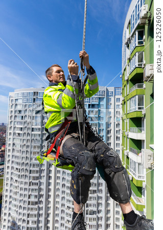 A Dedicated Worker Performs High-Rise Maintenance While Suspended in the Air, Secured by Ropes Near Tall Modern Apartment Buildings on a Clear Sunny Day 125226050