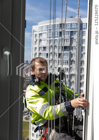 A Professional Window Cleaner Suspended at Height, Focused on His Task While Working on a Modern High-Rise Building in Bright Sunlight, Showcasing Urban Life and Safety 125226070