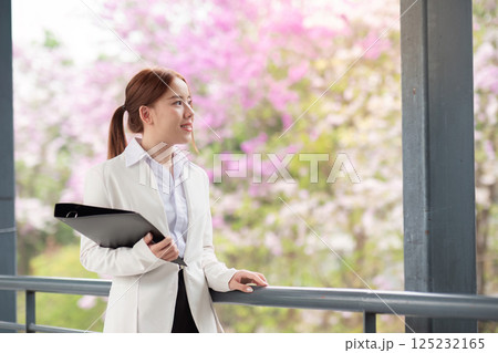Reflective businesswoman holding files and gazing at blooming flowers in a modern office setting 125232165