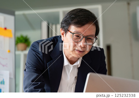 Elderly professional engaged in online work with a laptop and financial data. Elderly professional engaged in online work with a laptop and financial data. 125232338