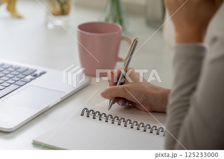 Writing and Productivity. A close-up of a woman jotting down notes while working on her laptop at home. 125233000